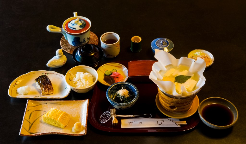 A Traditional breakfast at Tamahan Ryokan, Kyoto. The dishes include roast mackerel, dashimaki (Japanese omelette, here Kansai style), rice, a paper pot (kami nabe) of yudofu over a small brasier, with soy sauce and toppings (negi and katsuobushi), tsukemono (cucumber, pickling melon, turnip) and green tea. In the black cup is miso soup.
